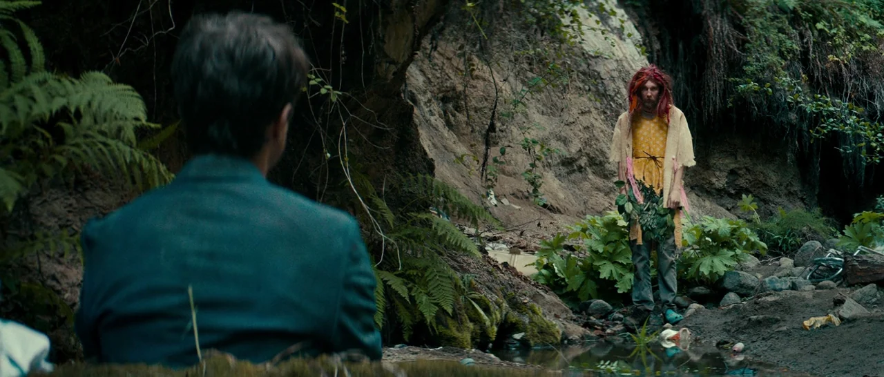 Swiss Army Man (2016) movie still: medium — A dark muddy ravine with thick vegetation and a narrow stream bed. A person in eccentric clothing stands…