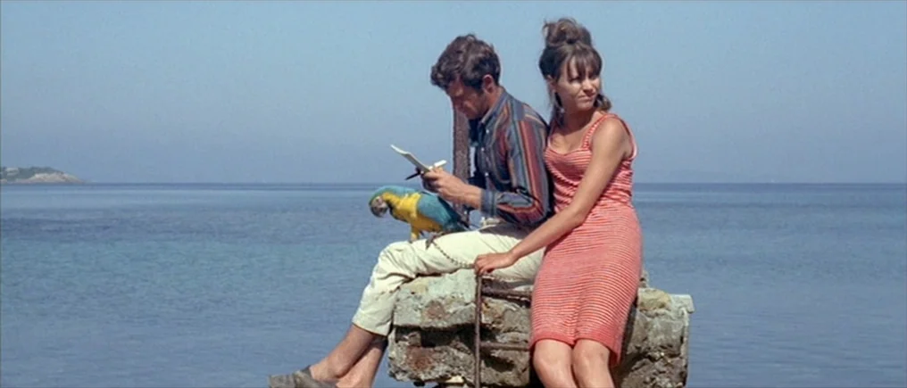 Pierrot Le Fou (Crazy Pete) (1965) movie still: fullbody — A man and woman sit together on a small stone structure surrounded by water. The man…