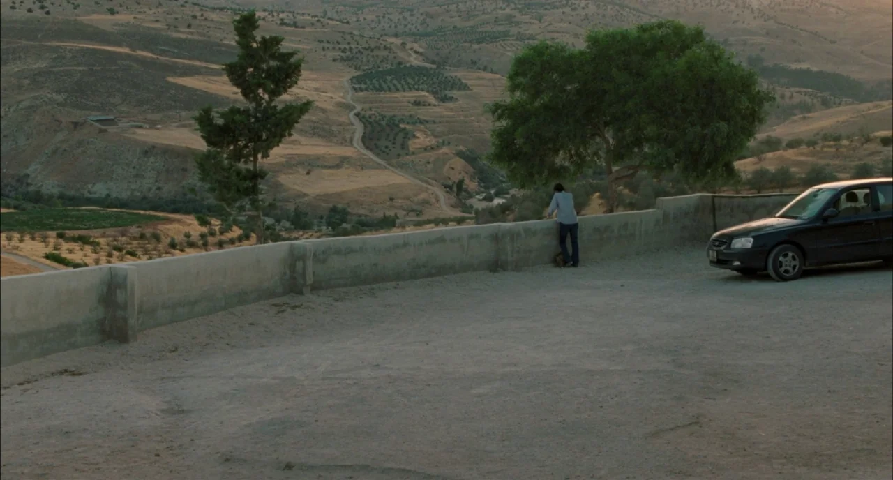 Incendies movie still: wide — A dry hilly landscape with sparse trees sits behind a low concrete retaining wall. A man stands at the wall…
