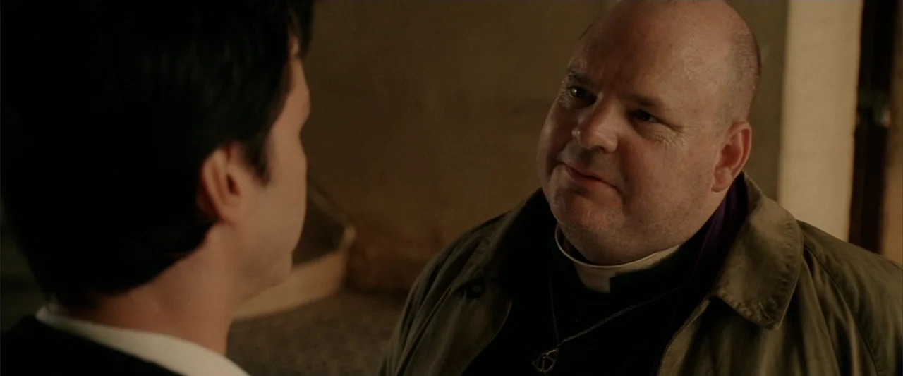 Constantine (2005) movie still: medium — A dimly lit indoor space with neutral-toned walls visible in the blurred background. A bald middle-aged man…