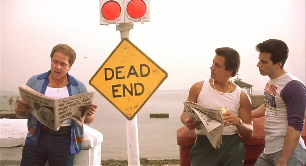 Summer of Sam (1999) movie still: medium — A group of three men stand next to a yellow traffic sign by the water. They hold newspapers while…