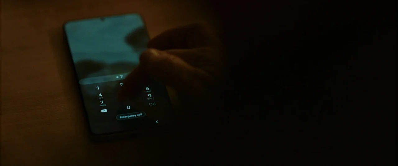My Son (2021) movie still: closeup — A dark, dimly lit room contains a smartphone resting on a wooden surface. A finger touches the digital keypad…