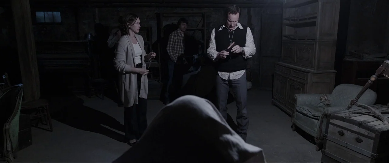 The Conjuring (2013) movie still: wide — A dimly lit basement features cluttered furniture including a piano, armchairs, and a hutch. A man in a…