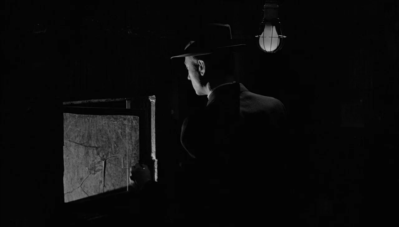 The Manchurian Candidate (1962) movie still: medium — A man in a fedora stands in a pitch-black room illuminated only by a bulb. He examines a small…