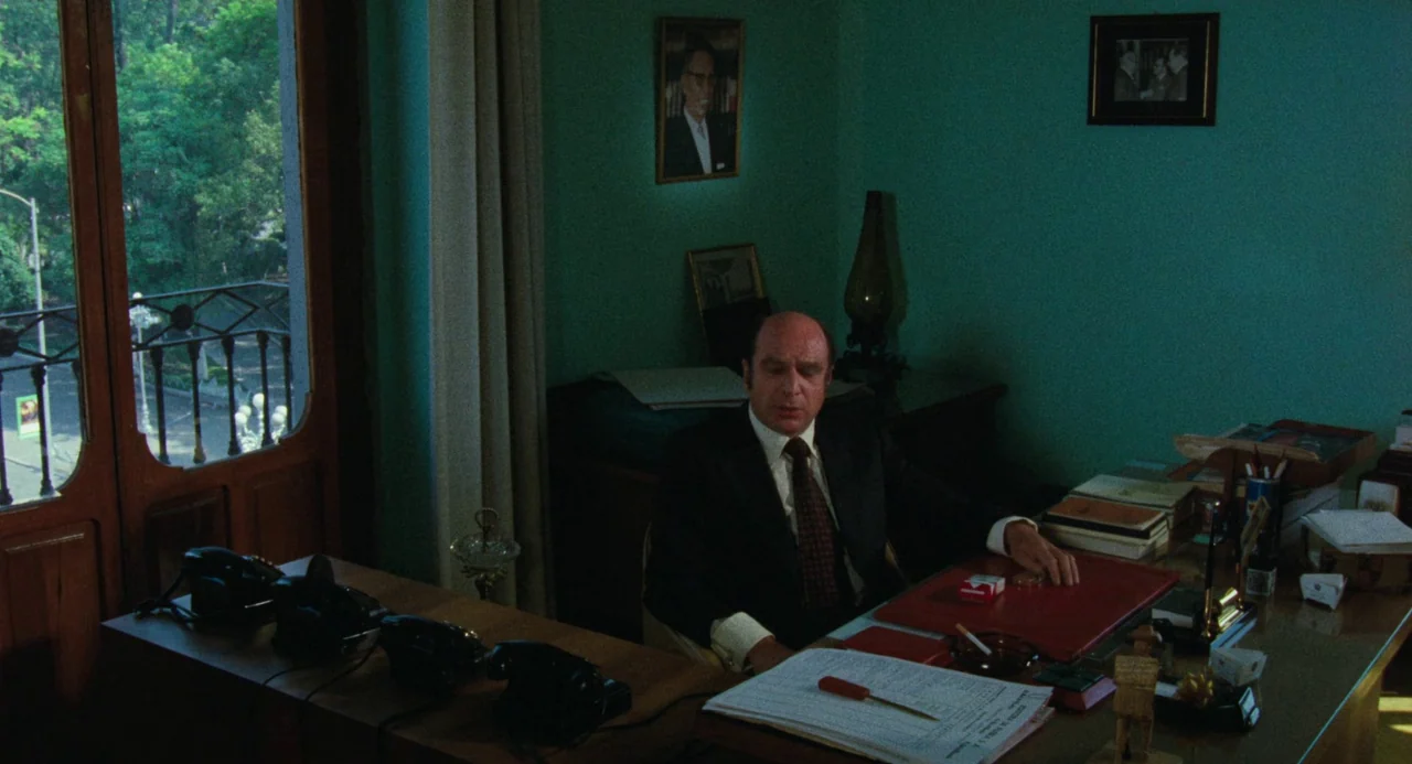 Canoa: A Shameful Memory (1976) movie still: medium — A teal-walled workspace features a wooden desk cluttered with paperwork and black telephones.…