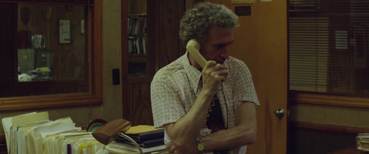 God’s Pocket (2014) movie still: medium — An aged 1970s workplace features wood-paneled walls and piles of paper files. A man in a patterned…