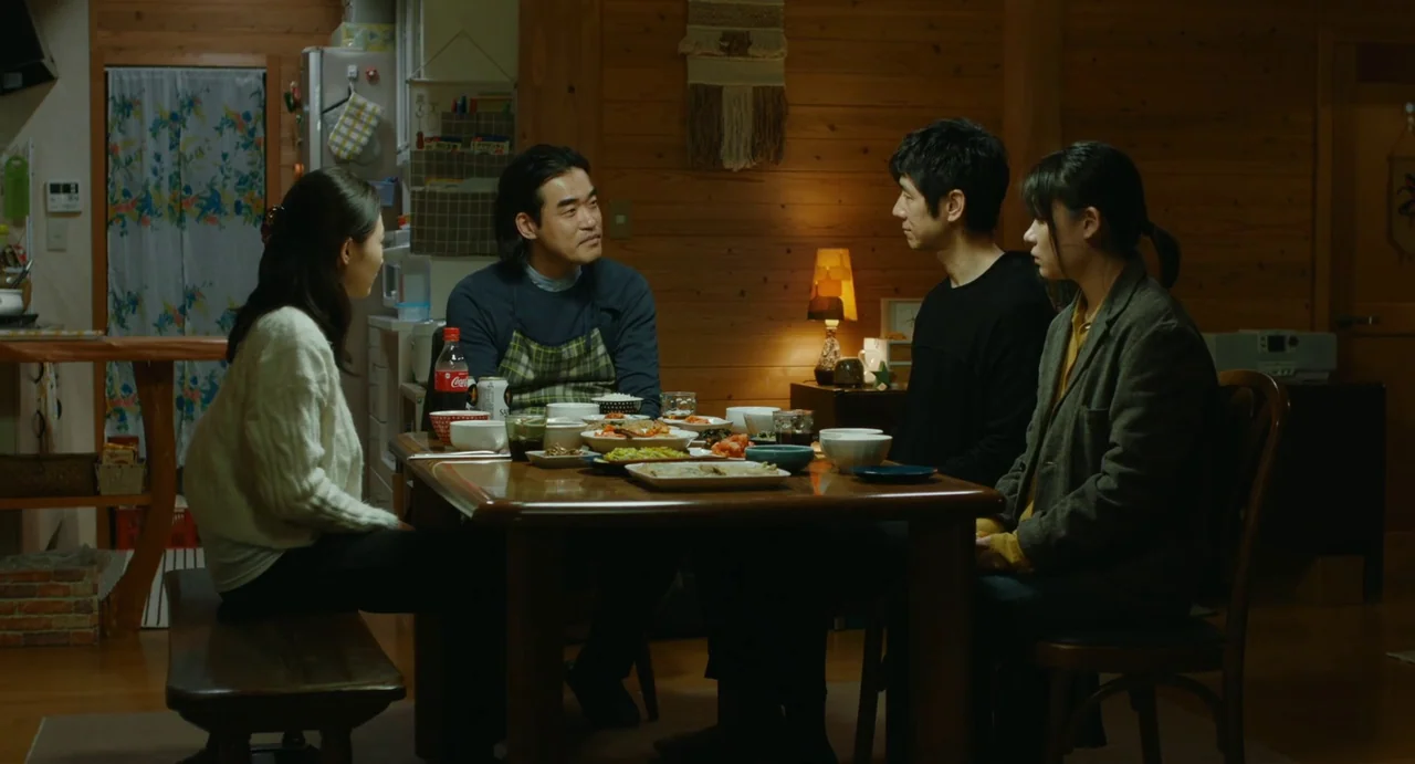 Drive My Car (2021) movie still: medium — A wooden paneled room features a dining table laden with dishes and shared food. Four adults sit around…