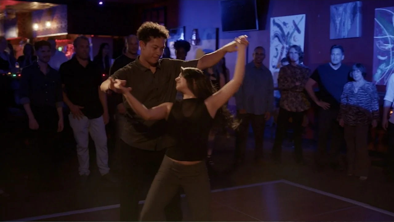 Ask Me to Dance (2022) movie still: medium — A dimly lit room features a hardwood dance floor surrounded by observers standing against walls. A…