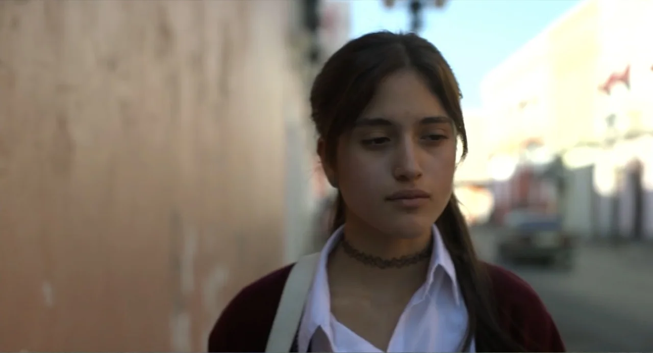 Heartbeat (2008) movie still: medium — A teenage girl walks along a paved pathway beside a textured peach-colored wall. The subject moves forward…