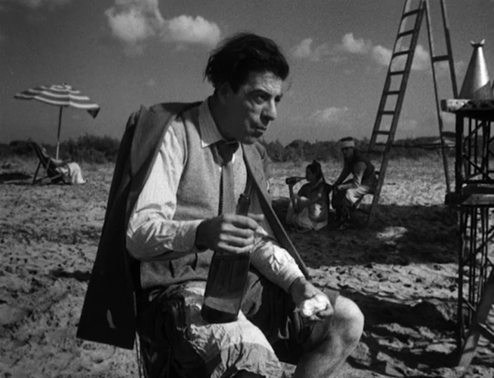 The White Sheik (1952) movie still: medium — A sandy shore contains a group of people sitting near a tall wooden ladder. The main man sits in the…