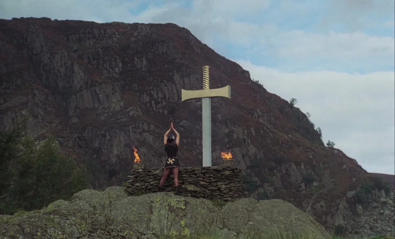 Mahler (1974) movie still: wide — A rugged mountainside features a dry stone wall platform supporting a large gold-hilted sword. A woman stands with…