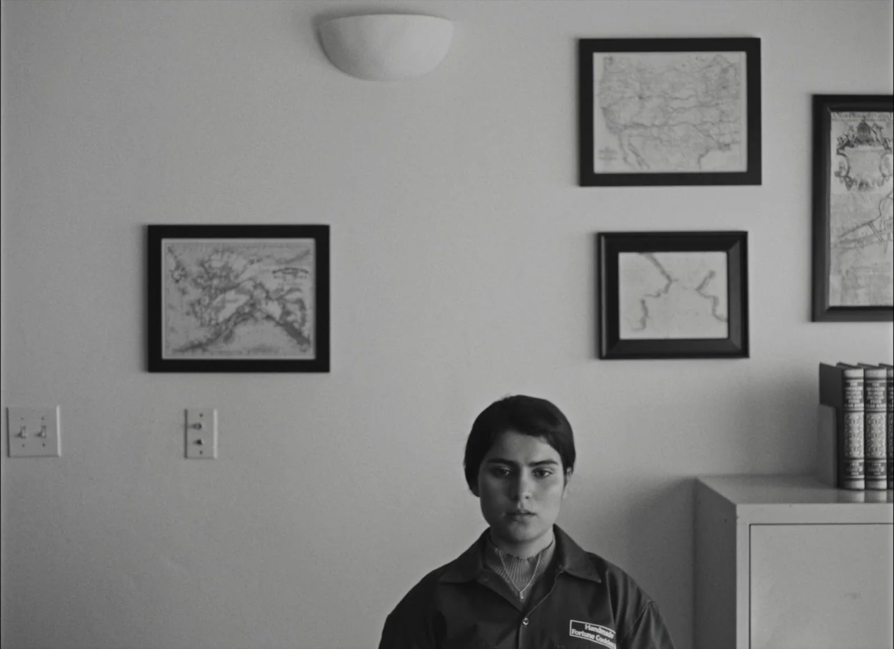 Fremont (2023) movie still: medium — A stark, minimalist white office wall features three framed maps and a light fixture. A young woman in a work…