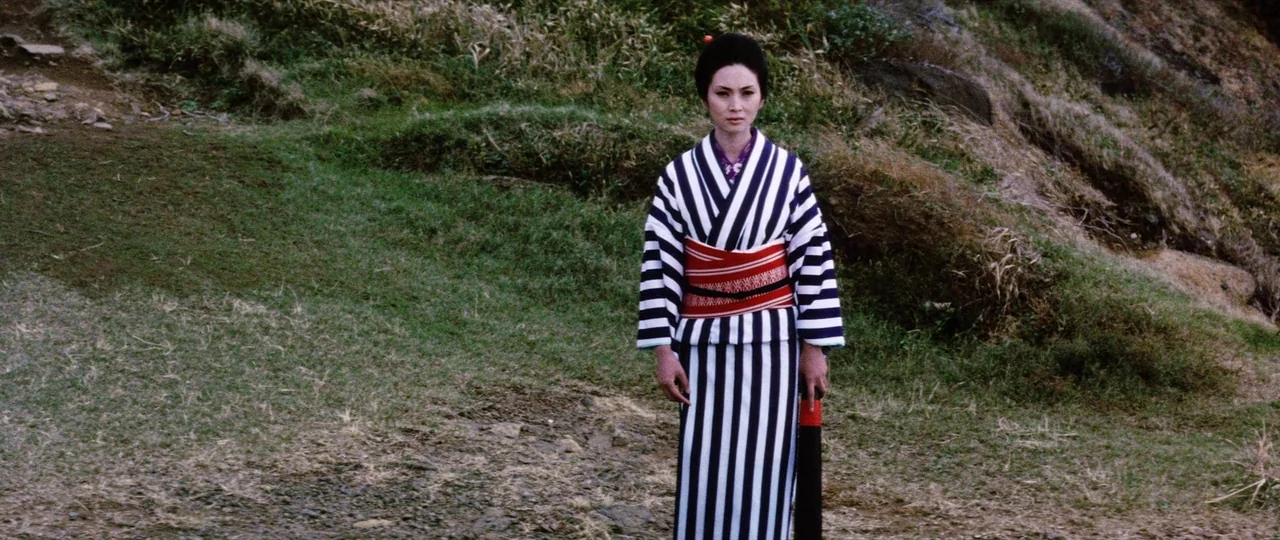 Lady Snowblood (1973) movie still: fullbody — A woman stands on a grassy hillside with a rock formation in the background. She wears a striped…