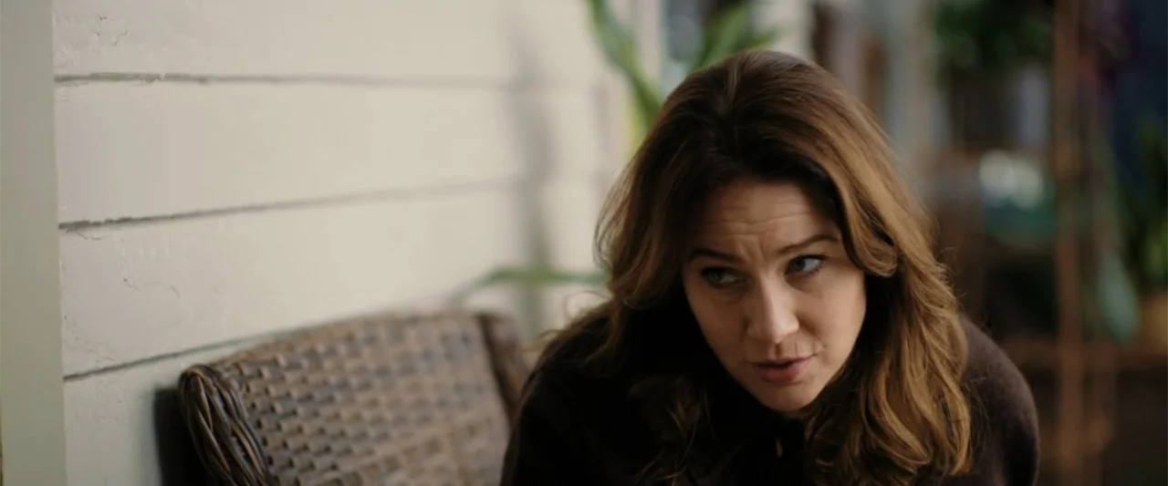 Someone Like You (2024) movie still: medium — A residential porch features white horizontal siding and a woven wicker chair. A woman sits on the…