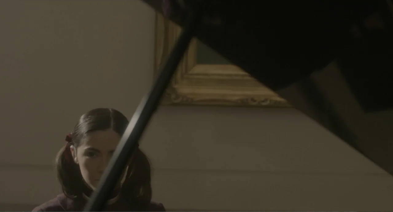 Orphan: First Kill (2022) movie still: medium — An interior space features a framed painting on a pale wall behind a piano. A young woman with her…