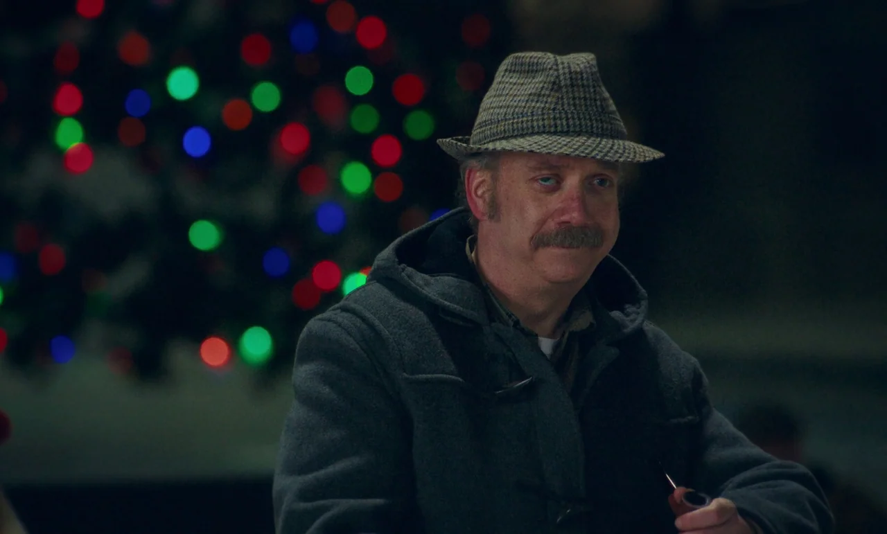 The Holdovers (2023) movie still: medium — A man stands in a dark school corridor with holiday light bokeh in the background. The man holds a wooden…