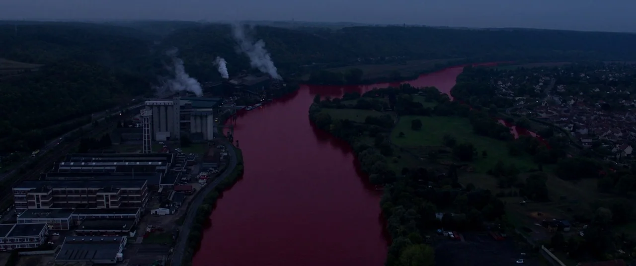 BPM (Beats Per Minute) (2017) movie still: establishing — An industrial facility with smoking chimneys sits beside a river turning a deep red. Green…