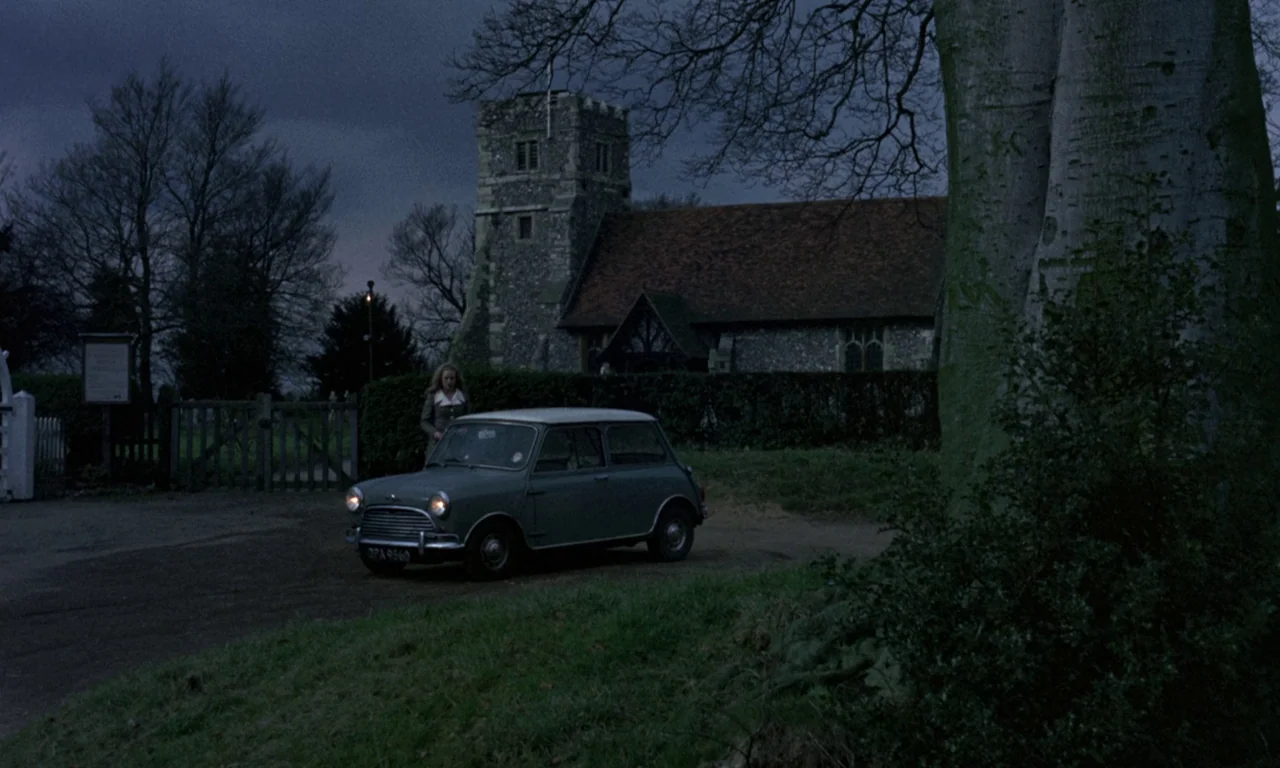 Curse of the Crimson Altar (1968) movie still: wide — A stone parish church sits behind a wooden gate under a darkening evening sky. A small green…