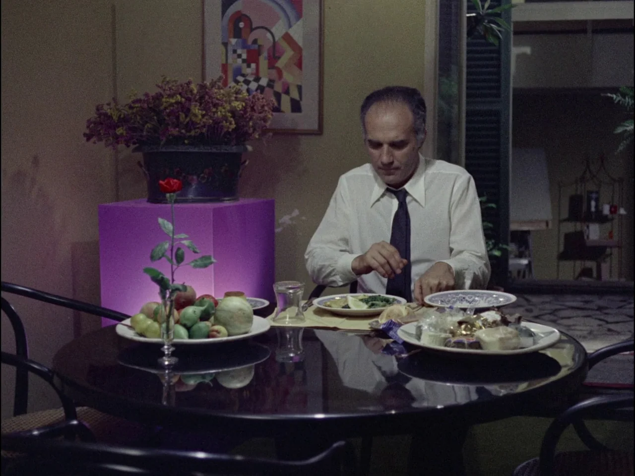 Dillinger is Dead (1969) movie still: medium — A man sits at a round glass dining table decorated with fruit and flowers. He eats a meal from a…