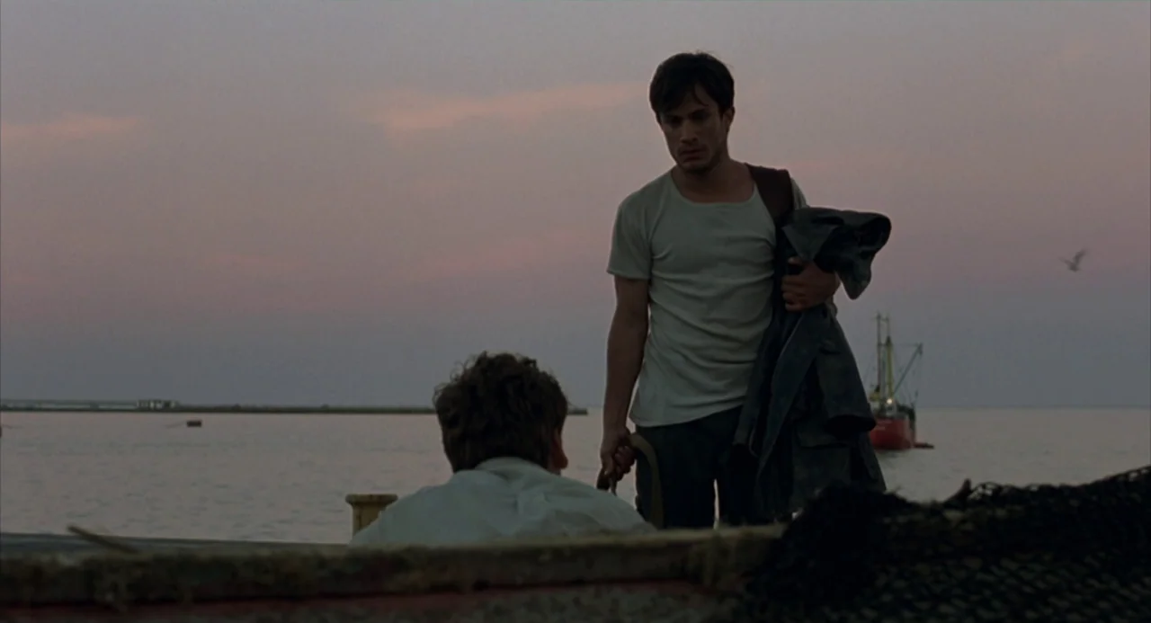 The Motorcycle Diaries (2004) movie still: medium — A wooden pier extends into the sea with a small fishing boat in the background. One man stands…