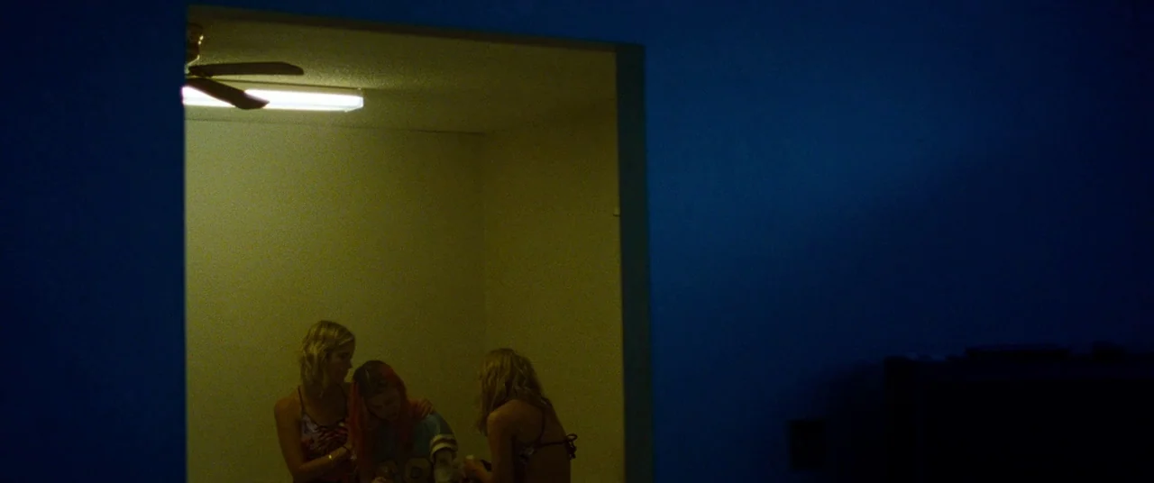 Spring Breakers (2012) movie still: wide — Three young women huddle together inside a small, brightly lit room. The surrounding space is dark,…