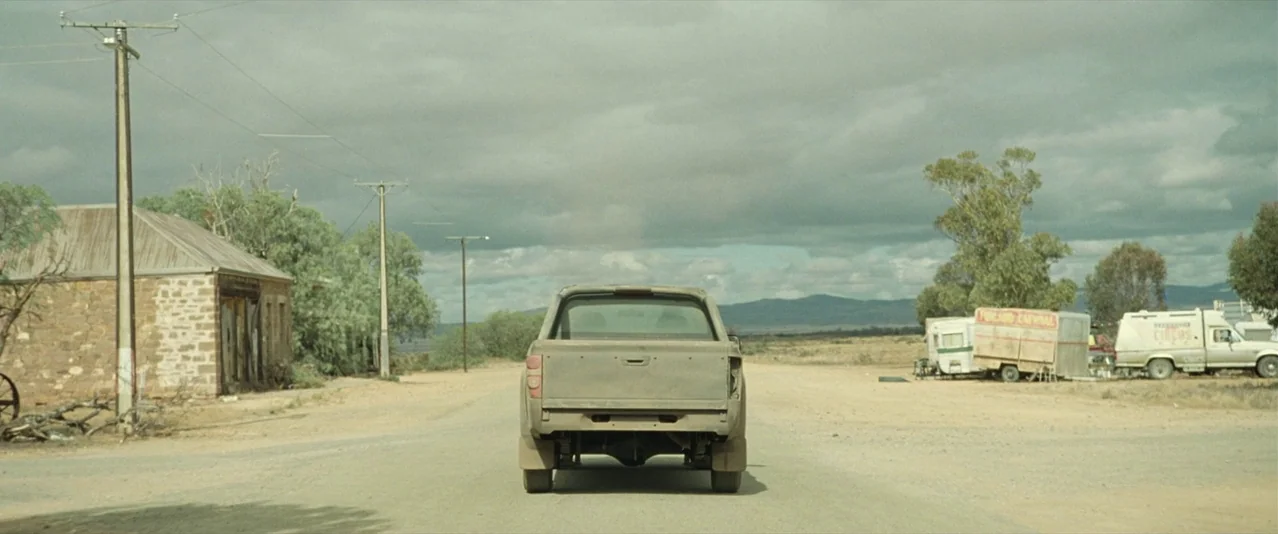 The Rover (2014) movie still: wide — A desolate rural road features a stone building on the left and trailers on right. A dusty, rear-facing pickup…
