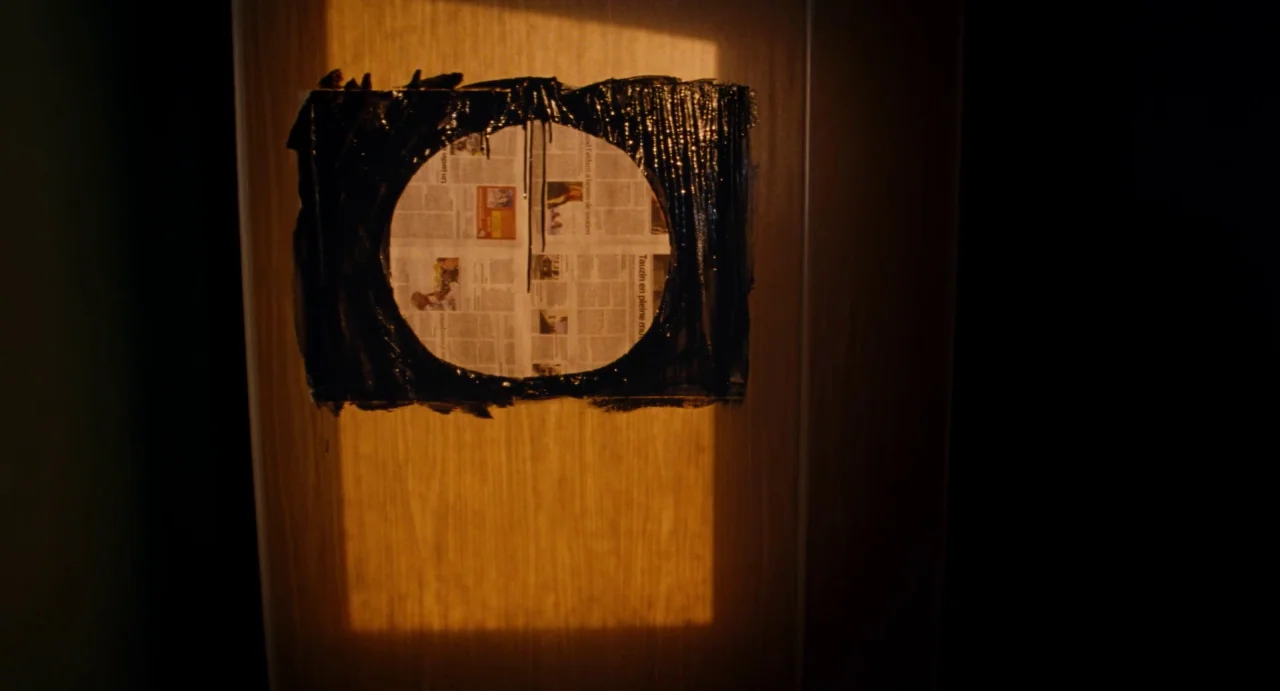 Ava (2017) movie still: closeup — A light-colored wooden door features a circular newspaper patch framed by black paint. A rectangular light pattern…