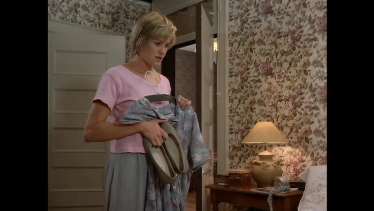 The Awakening (1995) movie still: medium — A bedroom features floral wallpaper and a wooden side table with a lamp. A young woman stands while…