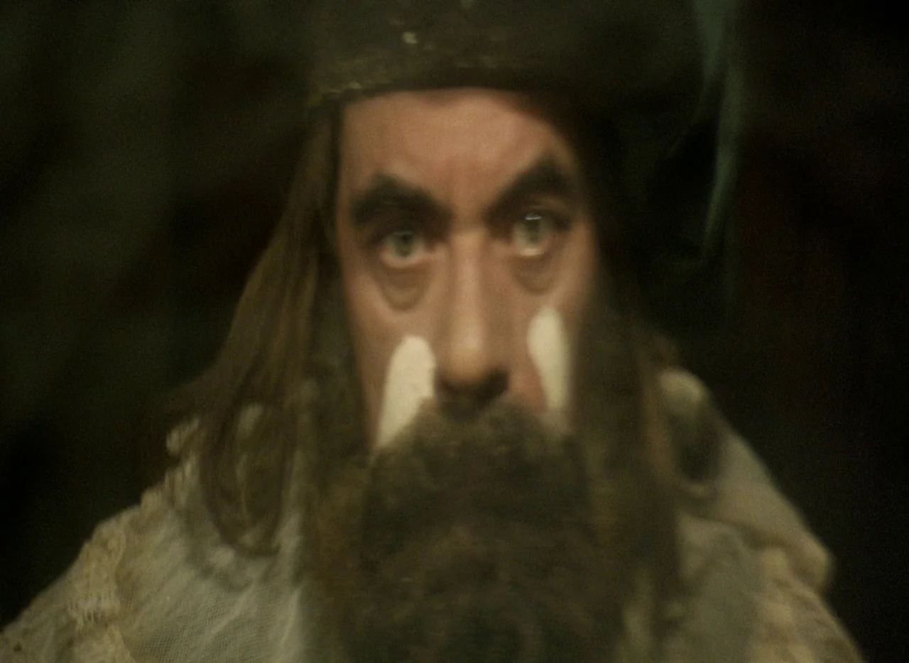Faust (1994) movie still: closeup — A dimly lit space with a dark background and indistinguishable surroundings. A man with face paint and a cap…