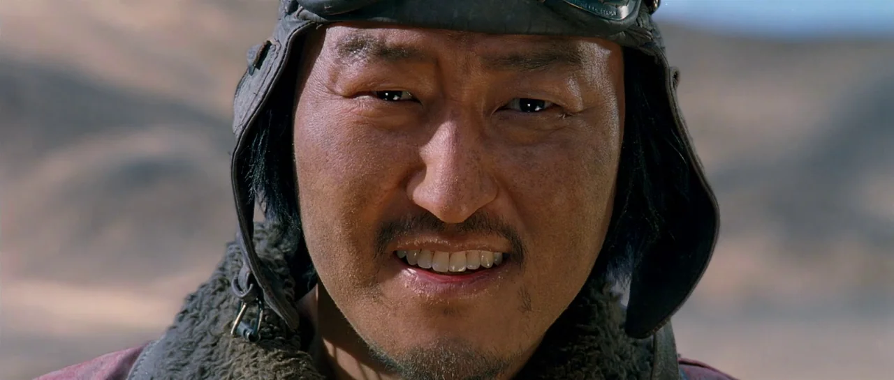 The Good The Bad The Weird (2008) movie still: closeup — An outdoor desert environment with a soft-focus background of sandy hills. A pilot in a…