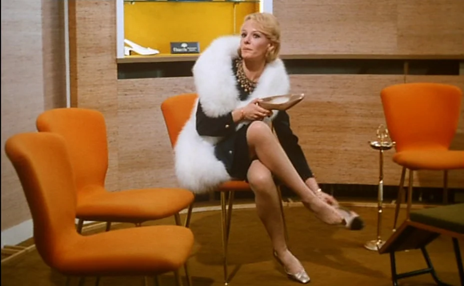 Stolen Kisses (1968) movie still: medium — A mid-century retail space with wood-paneled walls and bright orange modern furniture. A woman in a white…