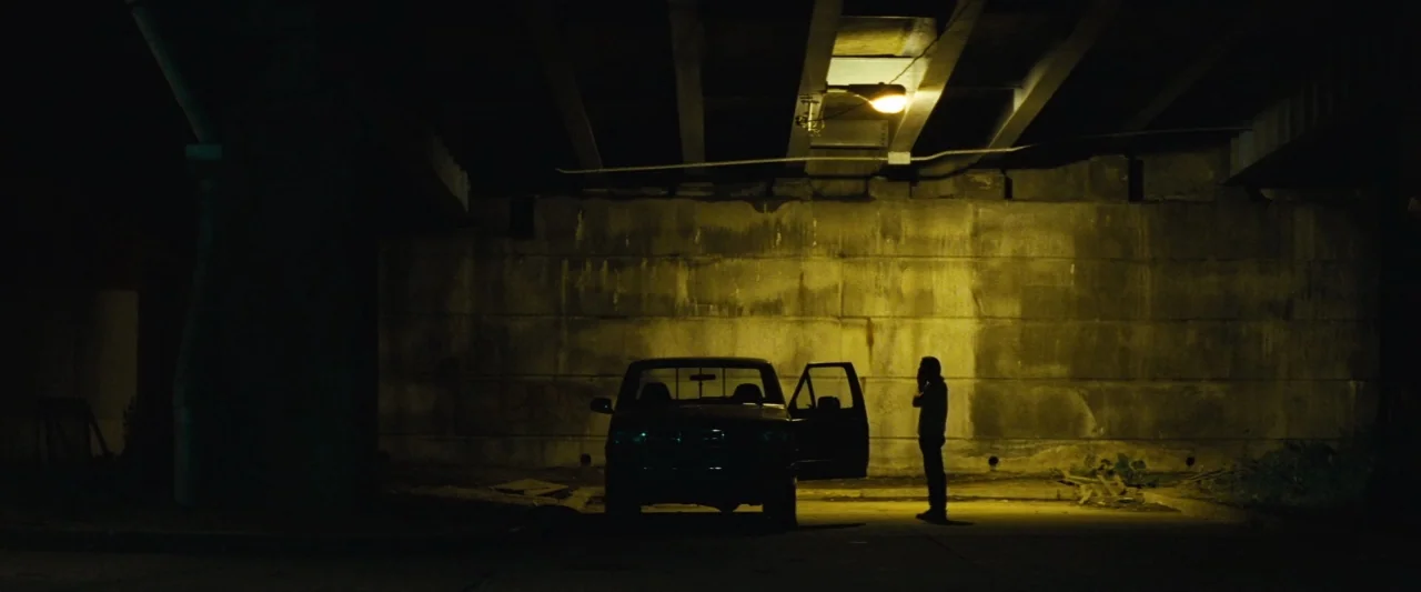 Out of the Furnace (2013) movie still: wide — A dark concrete underpass is lit by a harsh, single overhead yellow sodium lamp. A lone man stands…