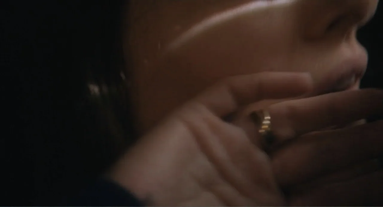May December (2023) movie still: closeup — A dark interior space is illuminated by a thin, sharp light source. A person's partial face and hand with…