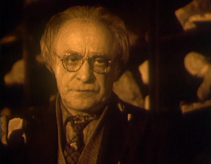 Dead Man’s Letters (1986) movie still: closeup — An elderly man is framed in a dark space filled with indistinct, pale objects. He stands directly…