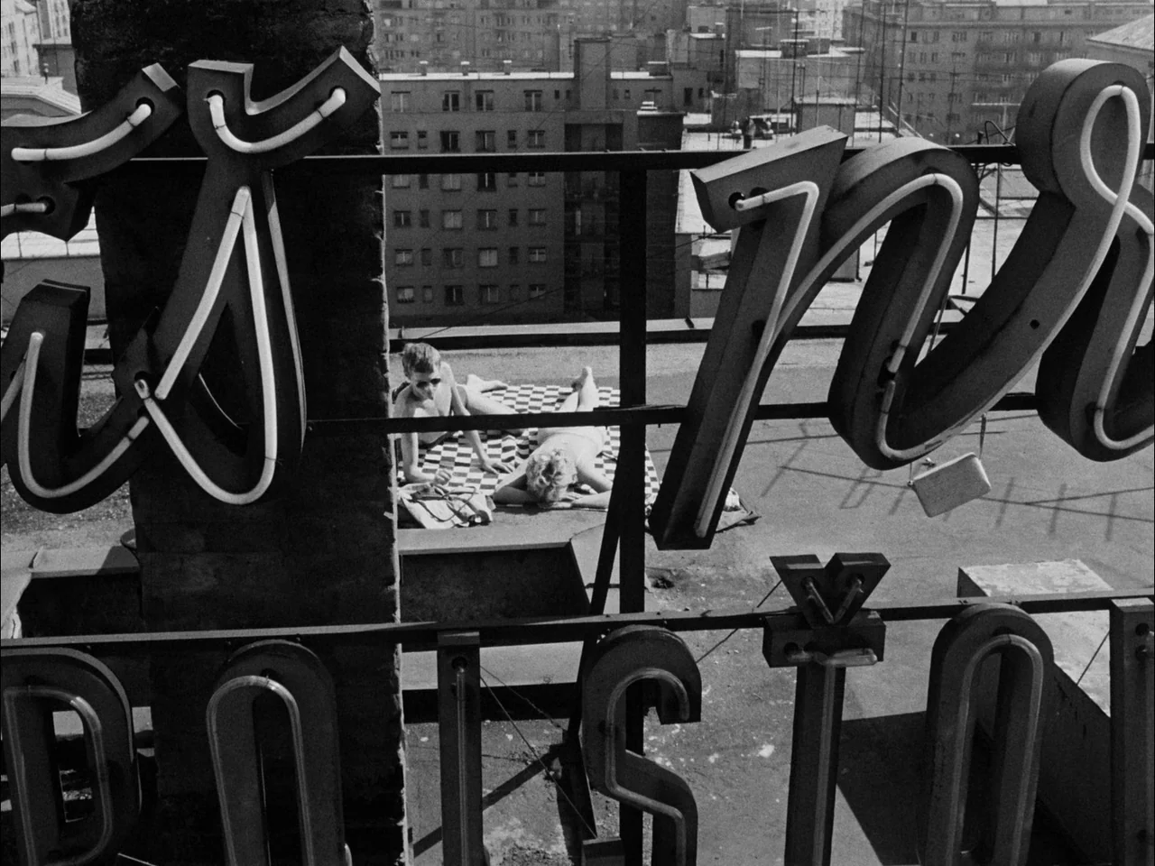 The Sun In A Net (1963) movie still: wide — A flat urban roof with a large metal neon sign structure in the foreground. A man and woman sunbathe on…