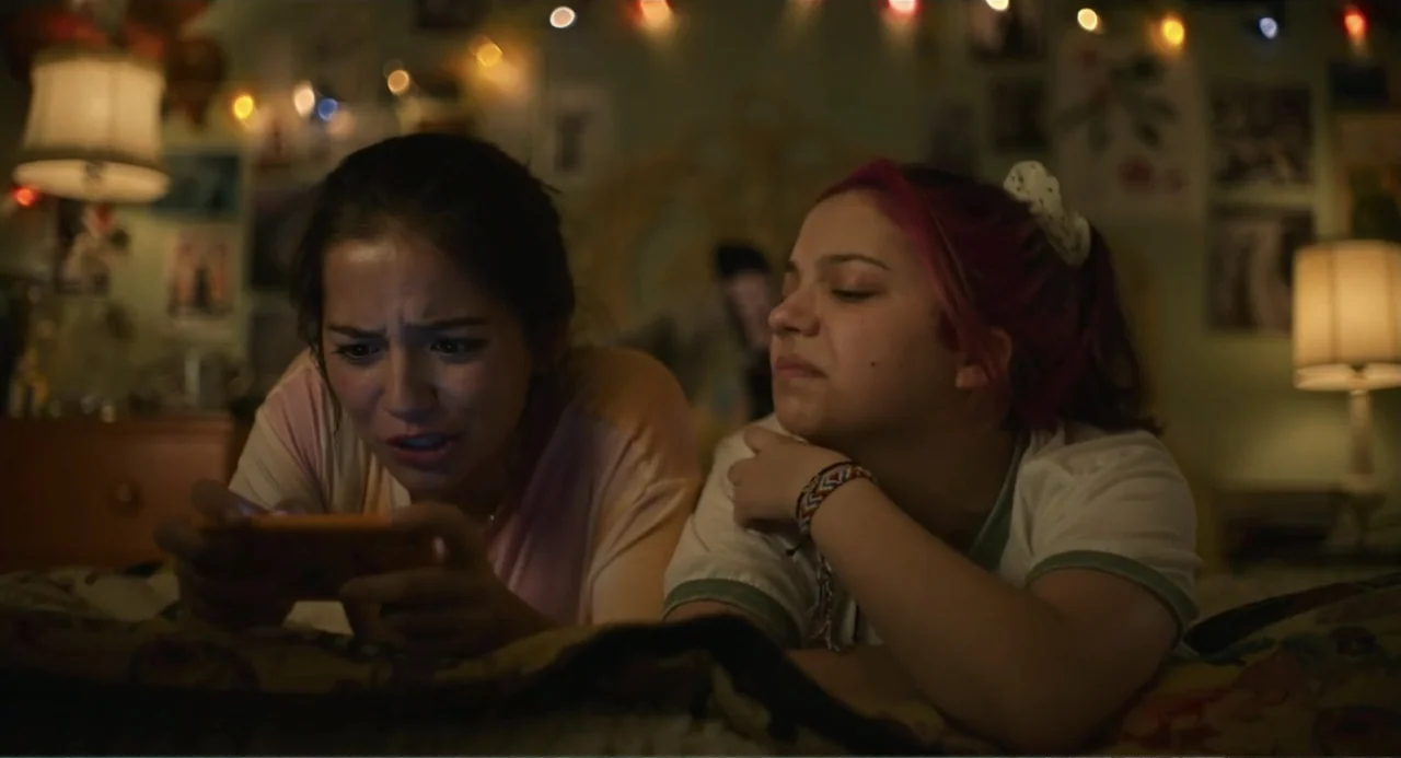Turtles All the Way Down (2024) movie still: medium — A dimly lit bedroom features string lights hung against a wall above a bed. Two teenage girls…