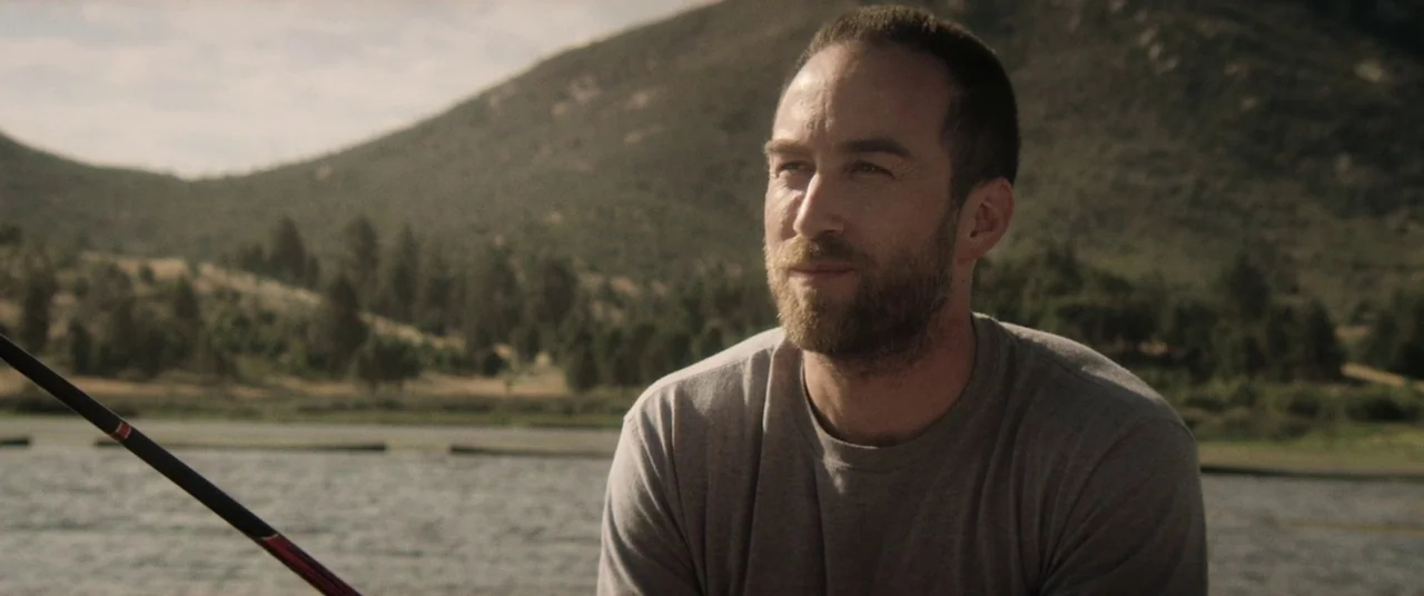 The Endless (2017) movie still: medium — A man sits by a lake with hills and trees visible in the background. He holds a fishing rod while looking…