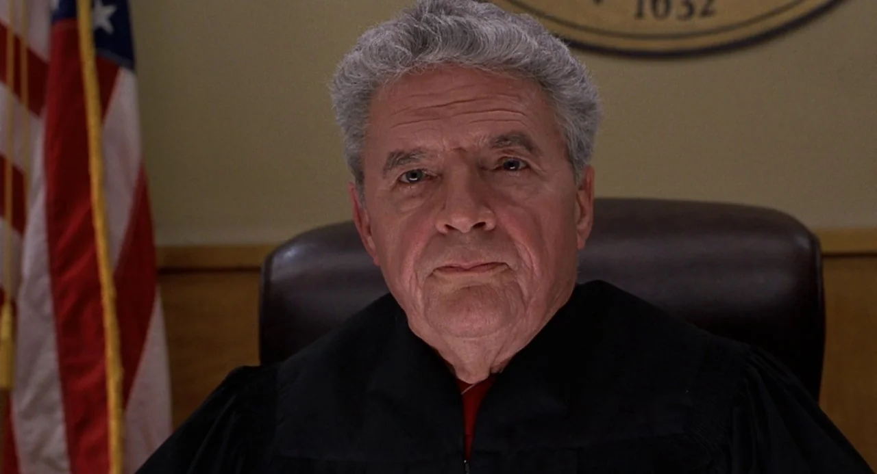 Cry Baby (1990) movie still: closeup — An elderly judge sits in a high-backed leather chair within a formal courtroom. A partially visible American…