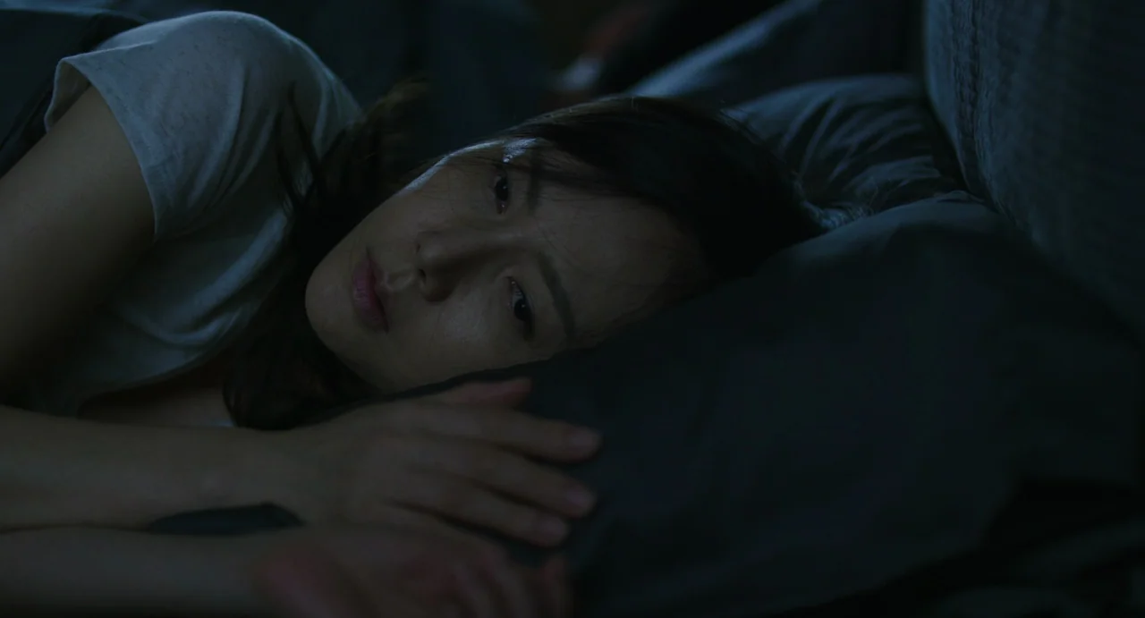 The Mimic (2017) movie still: closeup — A dark, dimly lit space with a person resting on a grey pillow. A woman lies in bed while staring straight…