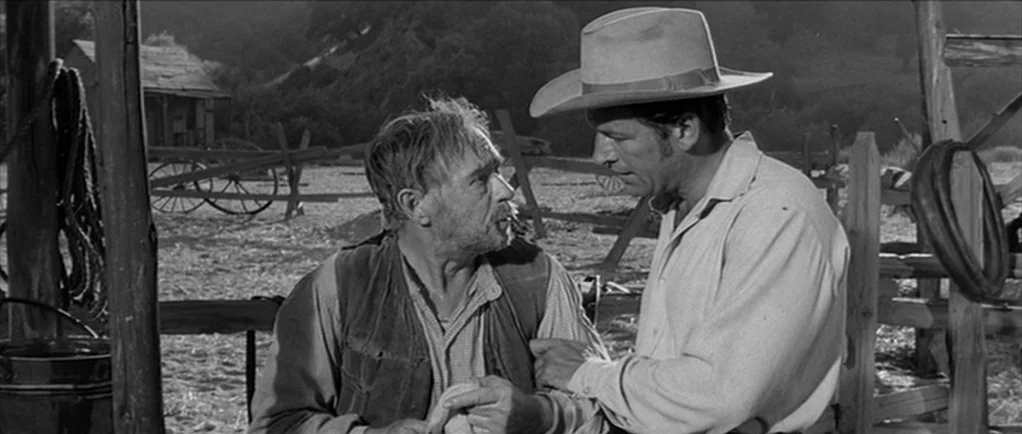 Love Me Tender movie still: medium — An outdoor ranch setting features a wooden fence and an old wagon in background. An older man seated is being…