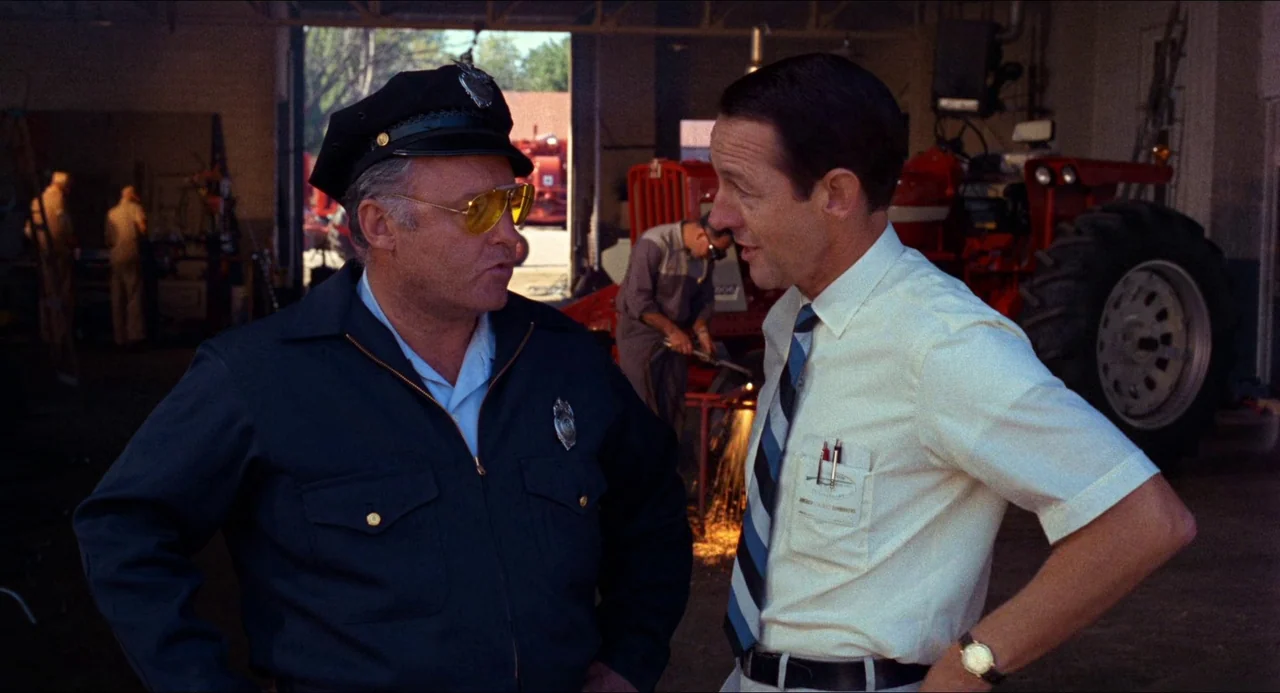 In the Heat of the Night (1967) movie still: medium — A spacious mechanic workshop contains a large red tractor and tools in the background. A…