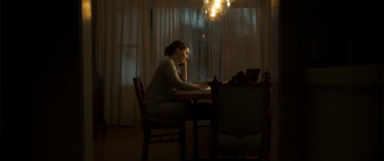 Phobias (2021) movie still: medium — A dark dining room features white curtains over a window and a wooden table. A woman sits alone at the table…