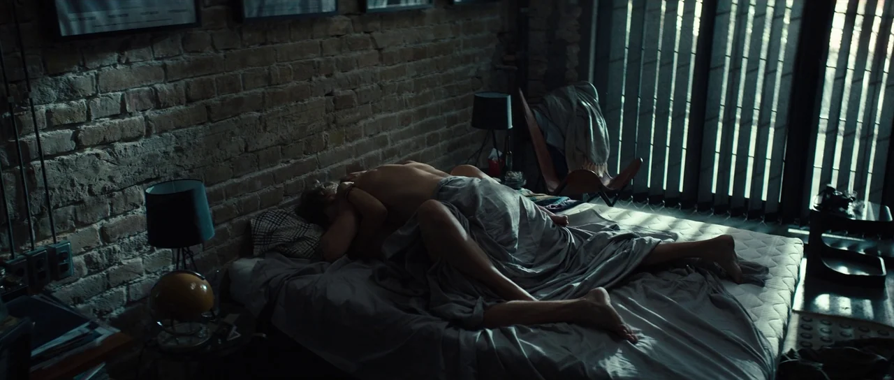 Hellraiser (2022) movie still: wide — A modern industrial bedroom features an exposed brick wall and a low platform bed. Two people are lying on the…