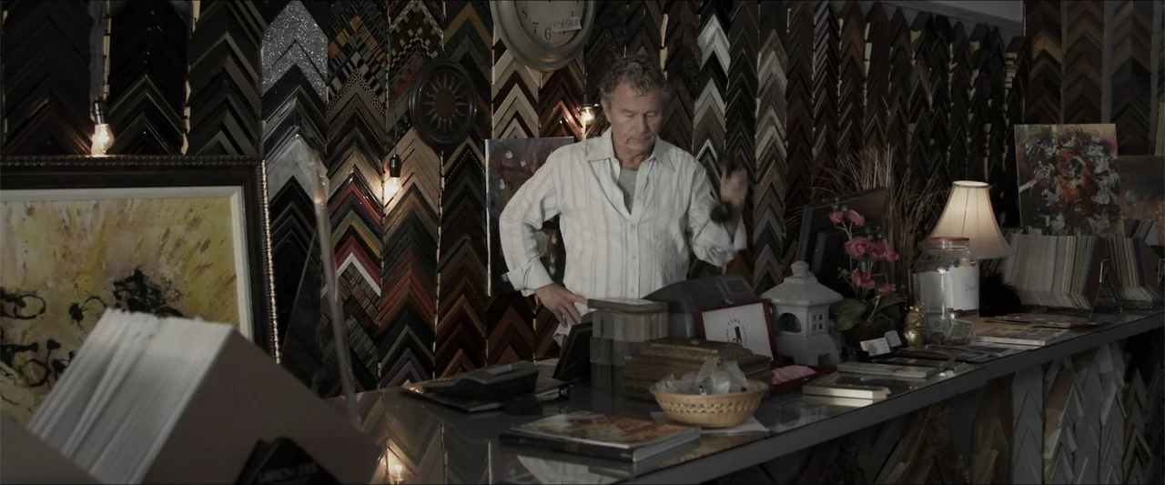 Heavenly Deposit (2019) movie still: medium — A store counter is covered in customer service items and stacks of framing samples. A man stands…
