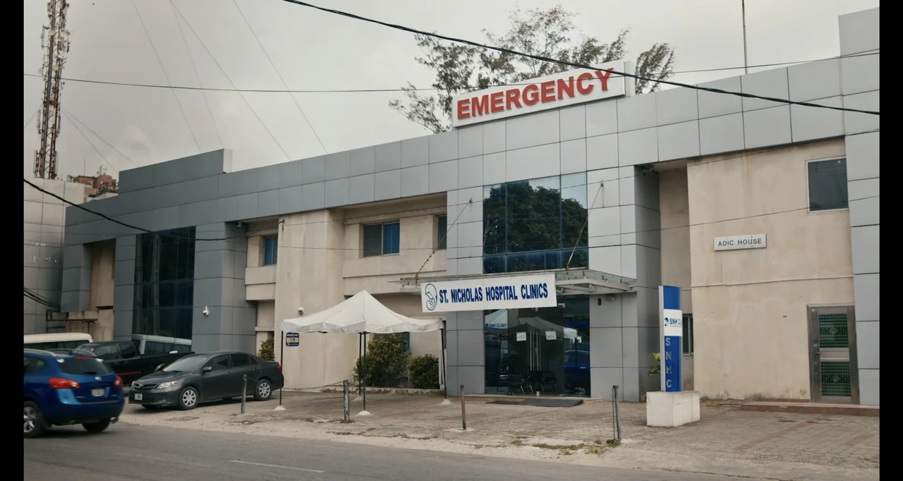 Love In A Pandemic (2023) movie still: wide — A modern clinic building with a grey panel facade and a prominent emergency sign. A parking area sits…