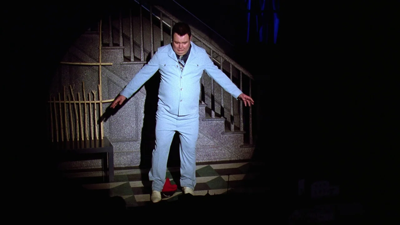Beetlejuice (1988) movie still: fullbody — A man stands on a stage floor with an staircase structure in the background. He is illuminated by a…