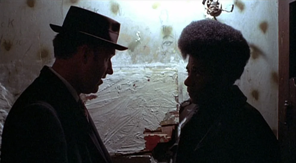 The French Connection (1971) movie still: medium — A small room features graffiti-covered walls and a damaged, textured patch of plaster. Two men…