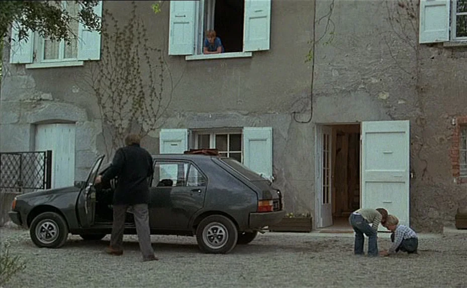 The Woman Next Door (1981) movie still: wide — A rural stone house with white window shutters sits behind a gravel parking area. A man climbs into a…