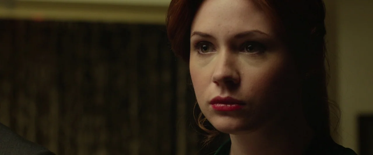 Oculus (2013) movie still: closeup — An interior space features a dark, textured background wall out of focus. A woman with red hair looks steadily…