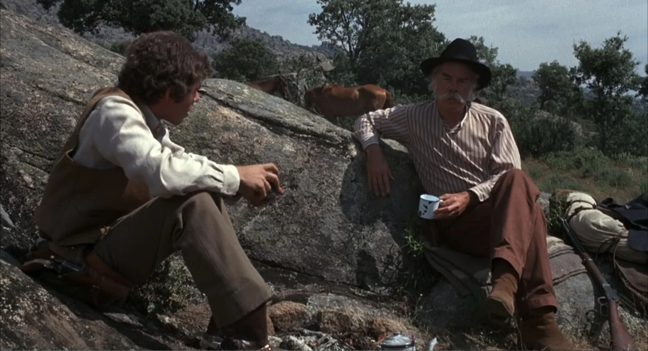 The Spikes Gang (1974) movie still: fullbody — A rocky hillside environment features large boulders, sparse shrubs, and grazing horses nearby. Two…